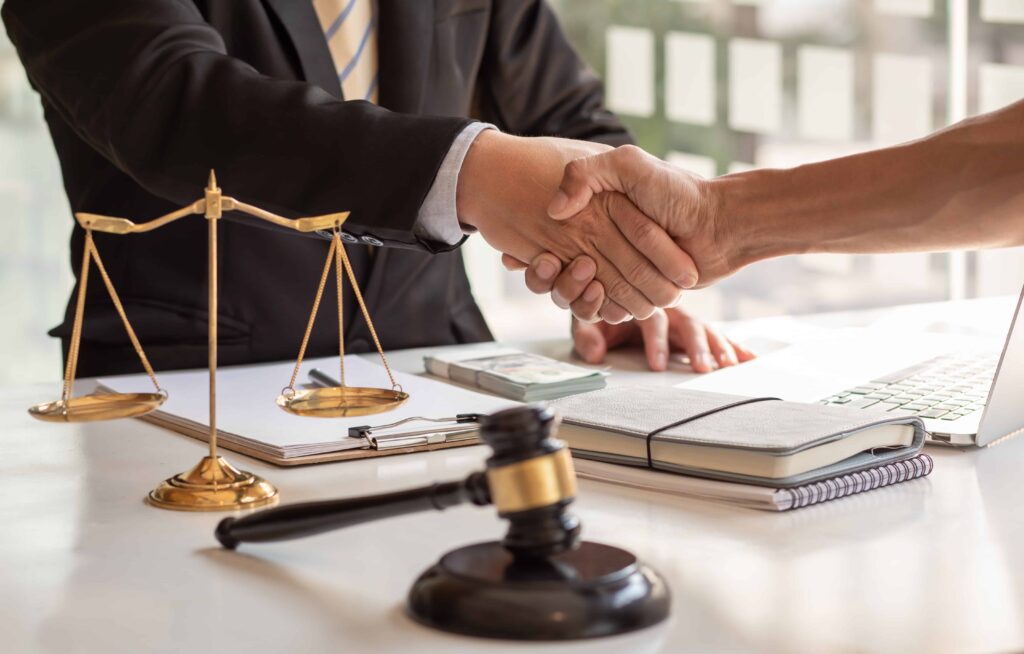 Lawyer and client shaking hands over legal documents with scales of justice and gavel on desk.
