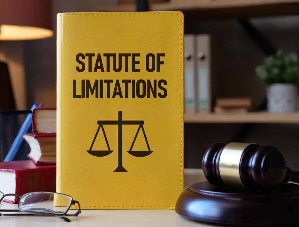 Yellow book titled ‘Statute of Limitations’ on a desk with a gavel, glasses, and legal books in the background.